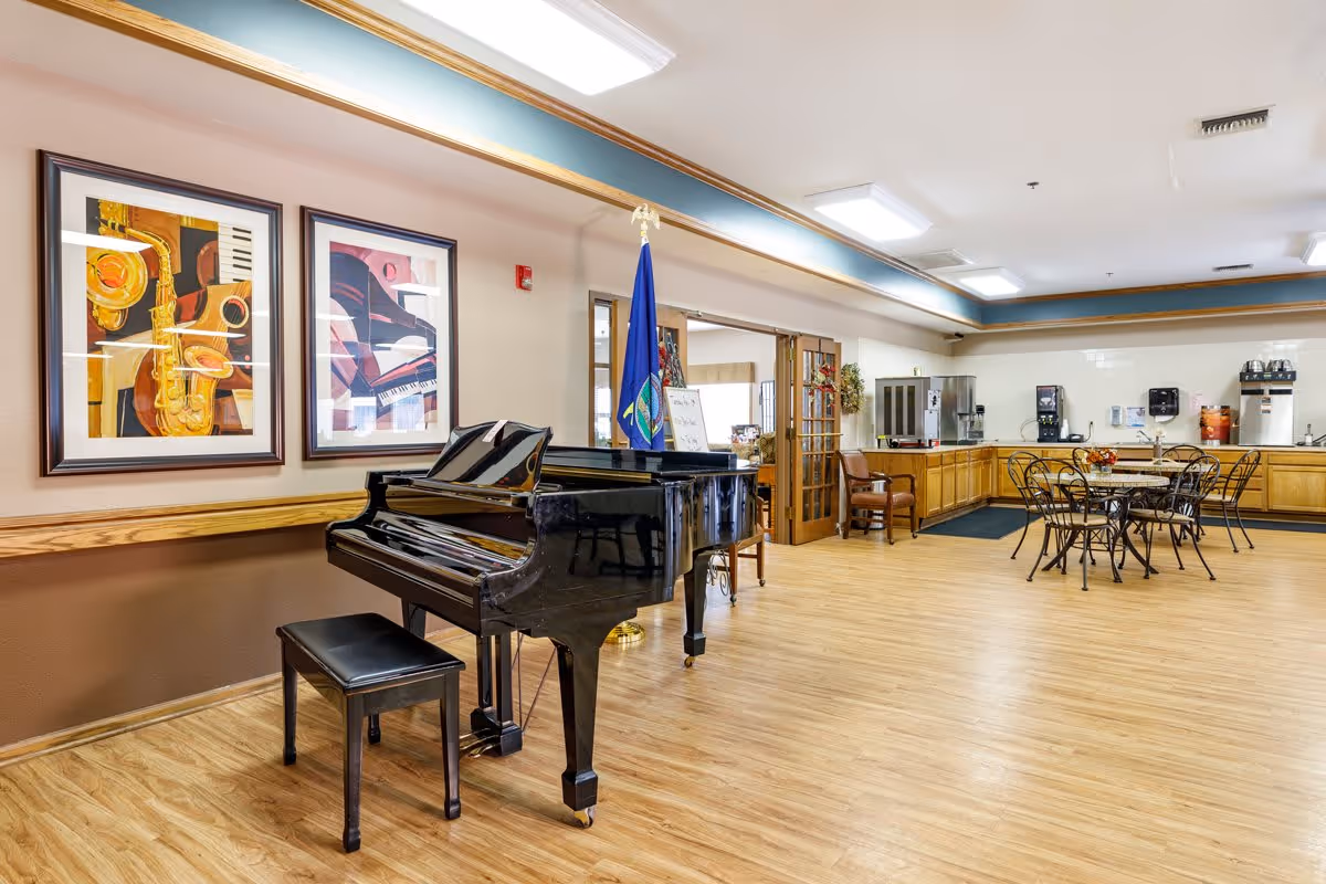 A spacious common area in a senior living facility featuring a black grand piano with a matching bench, two framed musical-themed paintings on the wall, a flag, and a seating area with round tables and chairs. The room has wood flooring, light-colored walls, and a counter with coffee and beverage machines in the background.