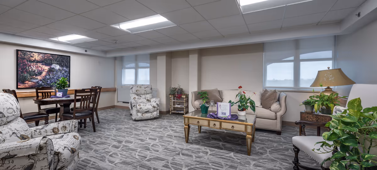 A spacious, well-lit living room area in a senior care facility featuring patterned armchairs, a beige sofa, a glass coffee table with plants and decorative items, a wooden dining table with chairs, and large windows with blinds. The room is decorated with indoor plants and a large framed painting on the wall.