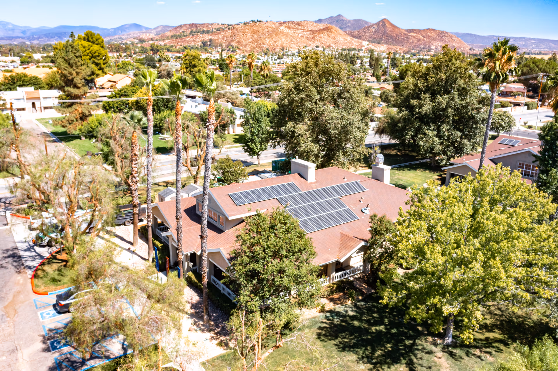 Aerial view of a single-story building with solar panels on the roof surrounded by trees, parking spaces, and distant mountains.