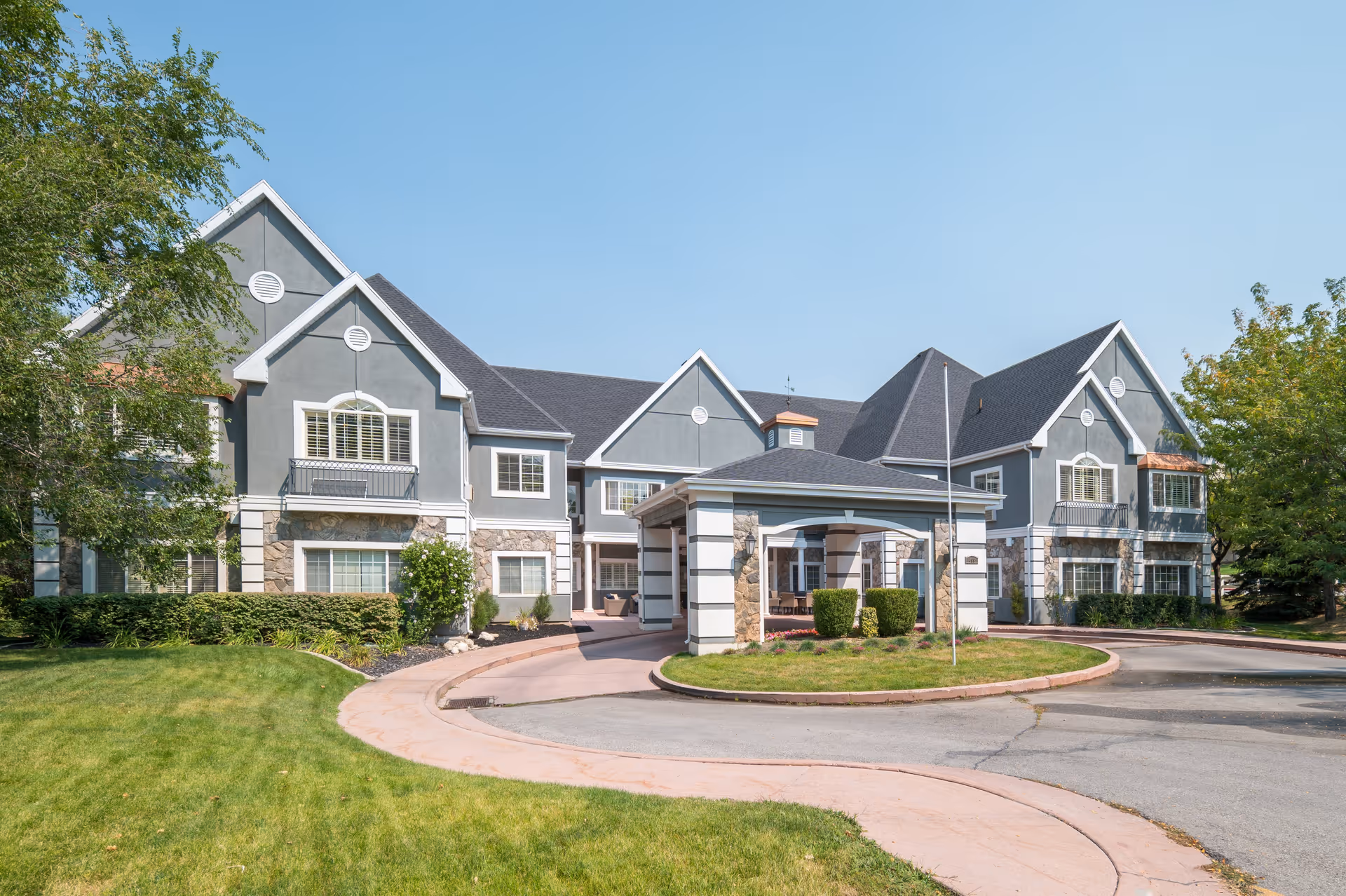 Exterior view of a senior living facility building with gray walls, white trim, and stone accents. The building has multiple peaked roofs and several windows with white shutters. There is a covered entrance with a circular driveway and well-maintained green lawns and trees surrounding the area under a clear blue sky.