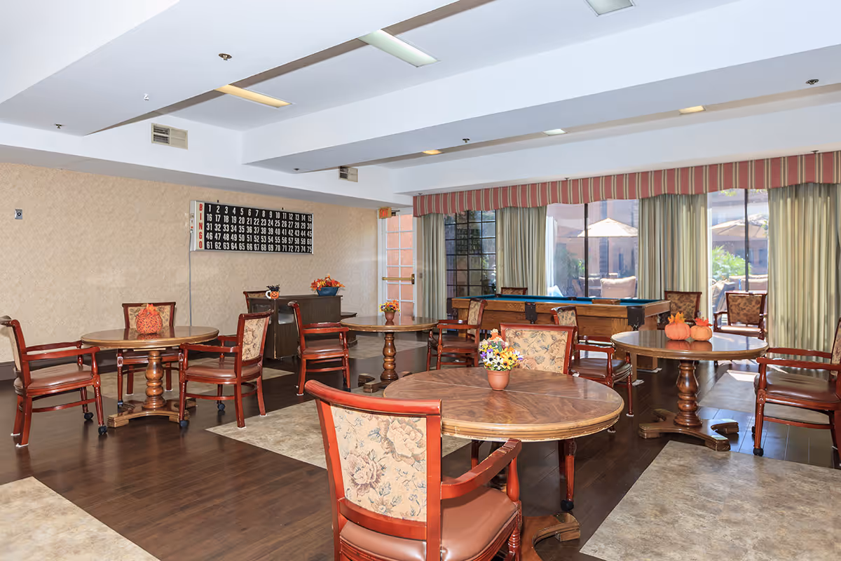 A spacious common room with several round wooden tables and cushioned chairs arranged around them. The room has large windows with curtains allowing natural light to enter, and a bingo board is mounted on the wall. There are small decorative items like flowers and pumpkins on the tables, and a pool table is visible near the windows.