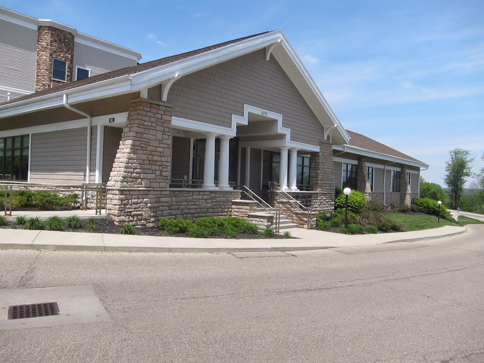Exterior view of a single-story building with stone and siding facade, featuring a covered entrance with white columns and steps leading up to the door. The building is surrounded by a paved driveway and landscaped greenery under a clear blue sky.