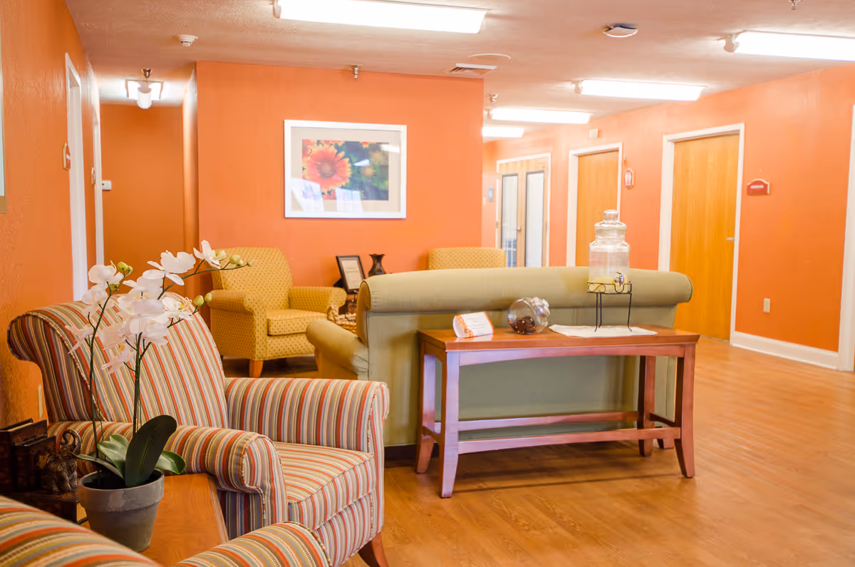A cozy interior common area with orange walls, wooden flooring, and several upholstered chairs and sofas arranged around a wooden table. A framed floral picture hangs on the wall, and a glass beverage dispenser sits on the table behind a green sofa.
