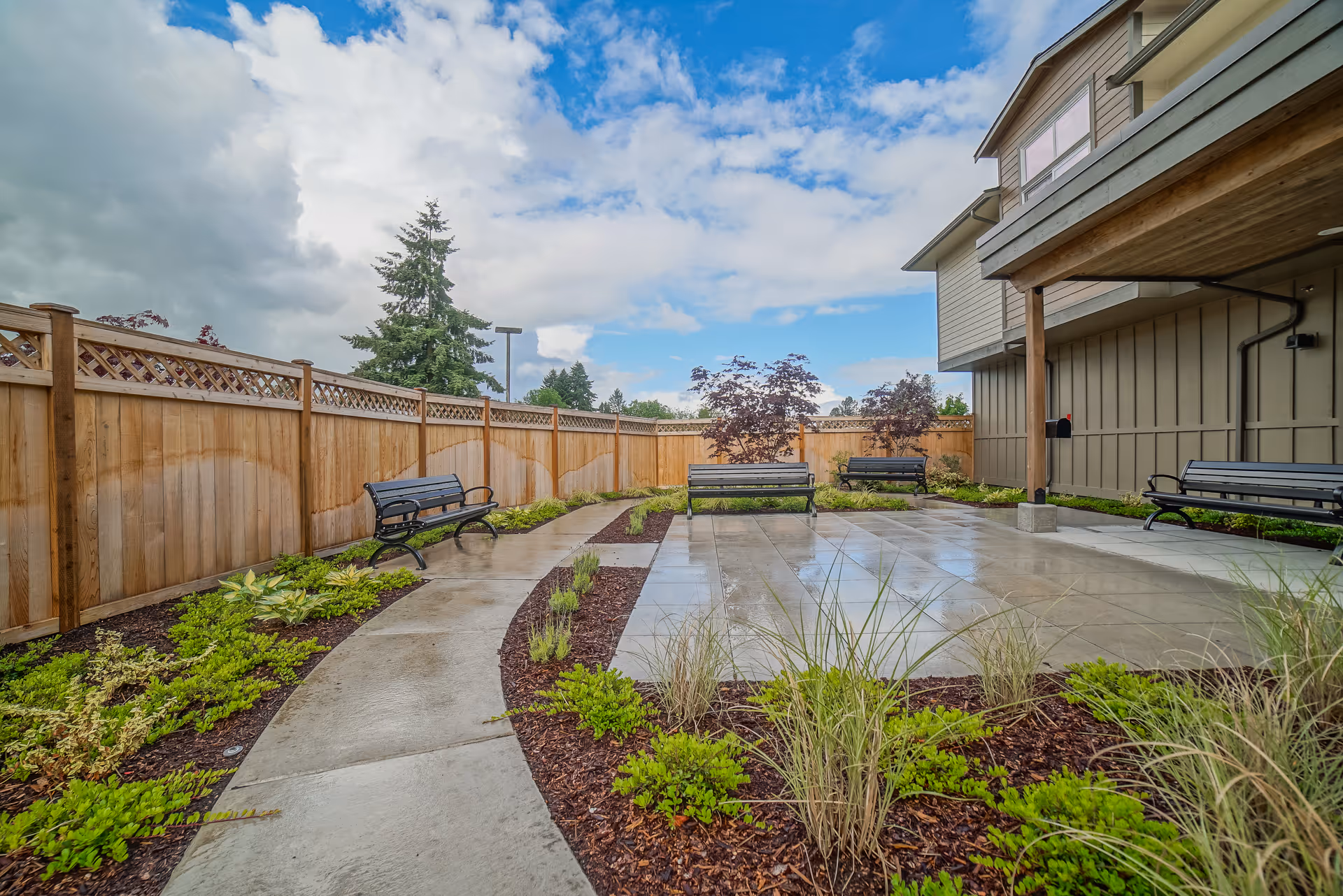 Patio courtyard with benches, paved walkway, and landscaped plant beds next to a building under a cloudy sky.