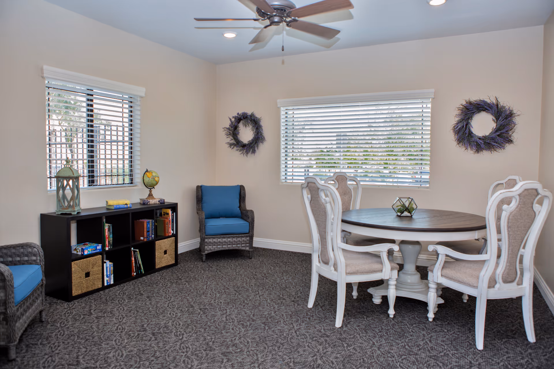 Bright common room with a round dining table and four chairs, wicker armchairs with blue cushions, a bookshelf, and windows with blinds.