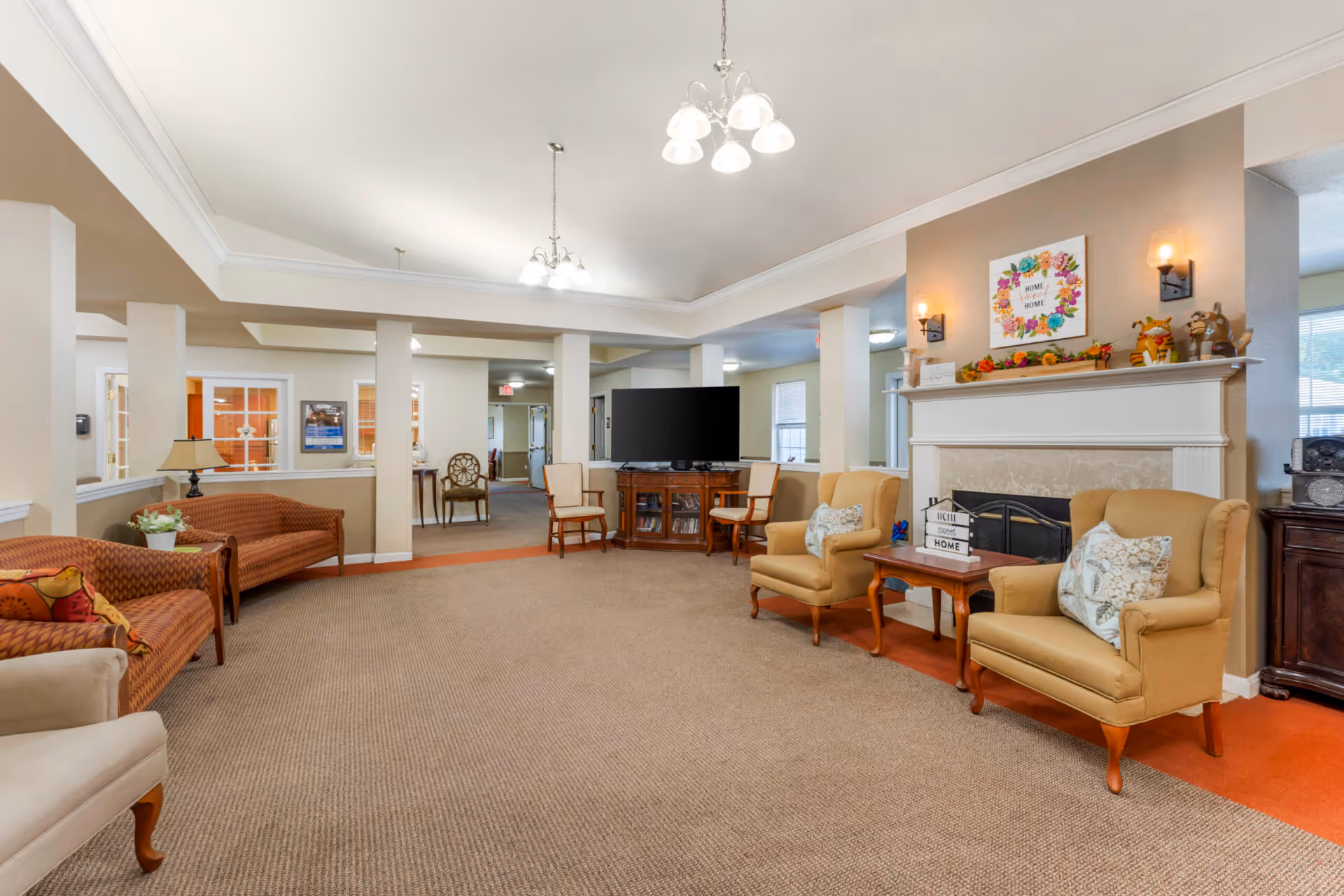 A spacious senior living facility common area with beige walls and carpeted floor. The room features comfortable seating including two yellow armchairs with patterned pillows, two brown patterned sofas, and wooden chairs. A fireplace with a mantel decorated with autumn-themed items and a framed 'Home Sweet Home' sign is on the right wall. A large flat-screen TV sits on a wooden stand in the background. The ceiling has white crown molding and two hanging light fixtures.