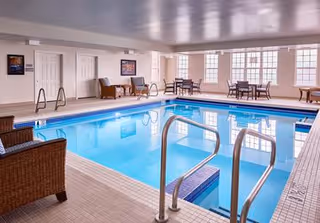 Indoor swimming pool area with clear blue water, metal handrails, wicker chairs, and tables with chairs near large windows allowing natural light to fill the space.