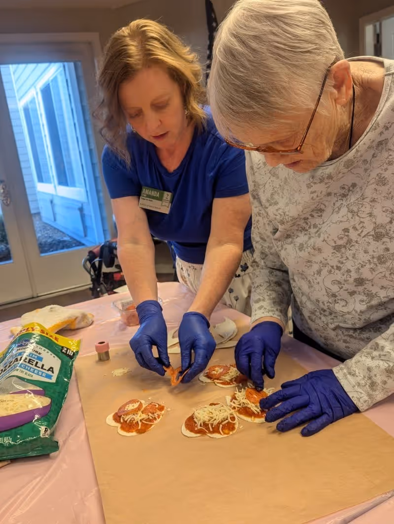 Two women wearing blue gloves are preparing mini pizzas on a table covered with brown parchment paper. One woman is placing pepperoni slices on the pizzas while the other is arranging toppings. A bag of shredded mozzarella cheese is visible on the table. The setting appears to be indoors with a door and window in the background.