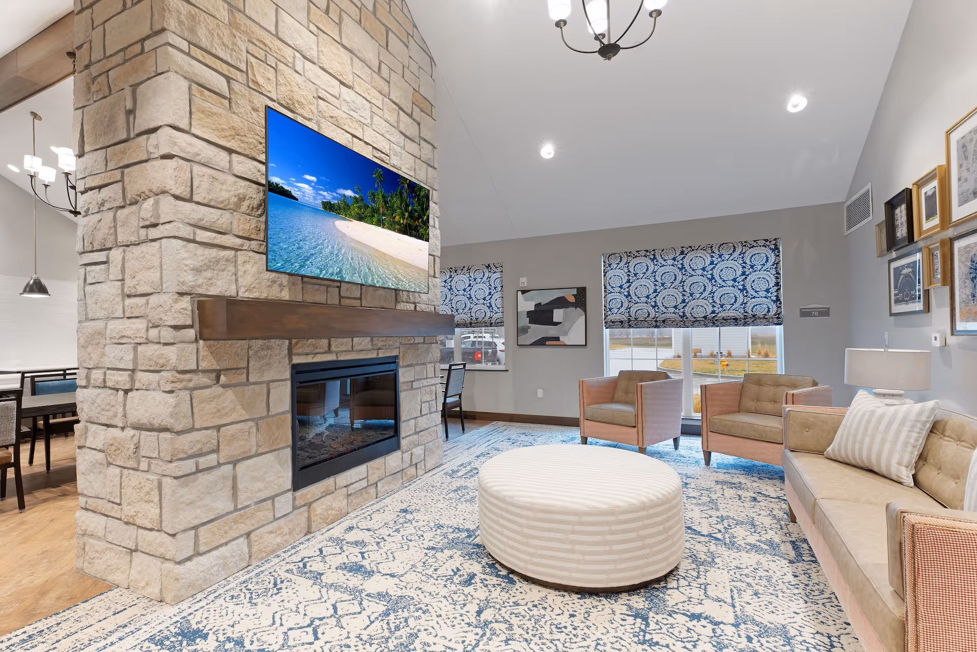A cozy senior living common area featuring a stone fireplace with a mounted flat-screen TV displaying a tropical beach scene. The room has a beige sofa with striped pillows, two armchairs, a round striped ottoman, and a blue patterned rug. Two windows with blue and white patterned Roman shades let in natural light. The walls are decorated with framed artwork, and a dining area is partially visible in the background.