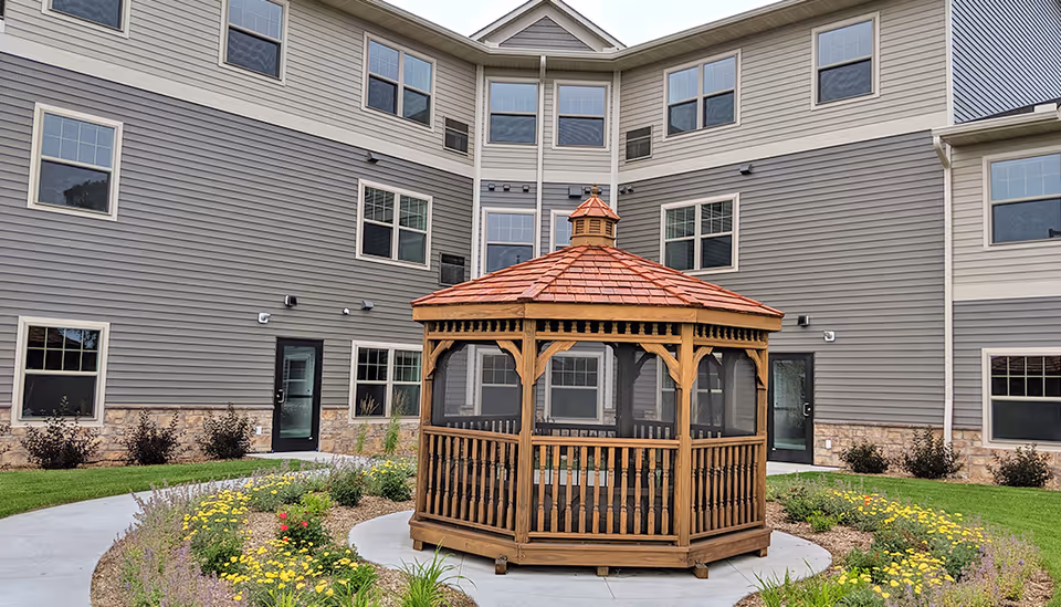 Wooden octagonal gazebo on a circular concrete pad surrounded by flower beds in a courtyard before a multi-story building.