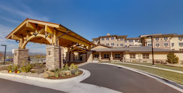 Exterior view of Wind Crest Senior Living Community building with a covered entrance supported by stone pillars, a driveway, landscaped plants, and a clear blue sky.