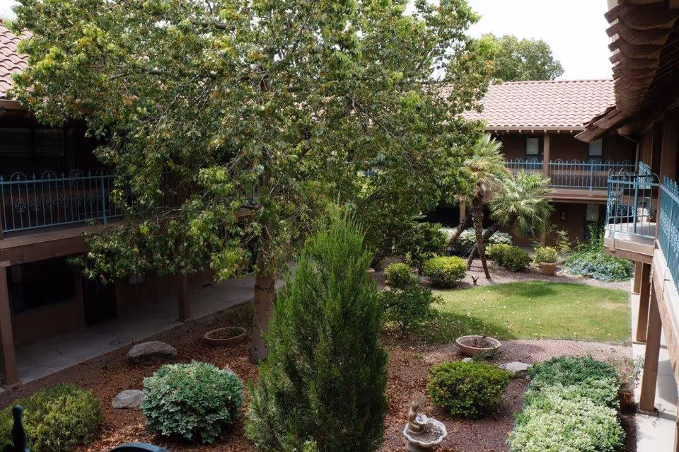 View of a landscaped courtyard garden in a senior living facility with various green shrubs, trees, and a small fountain. The courtyard is surrounded by a two-story building with balconies and railings.