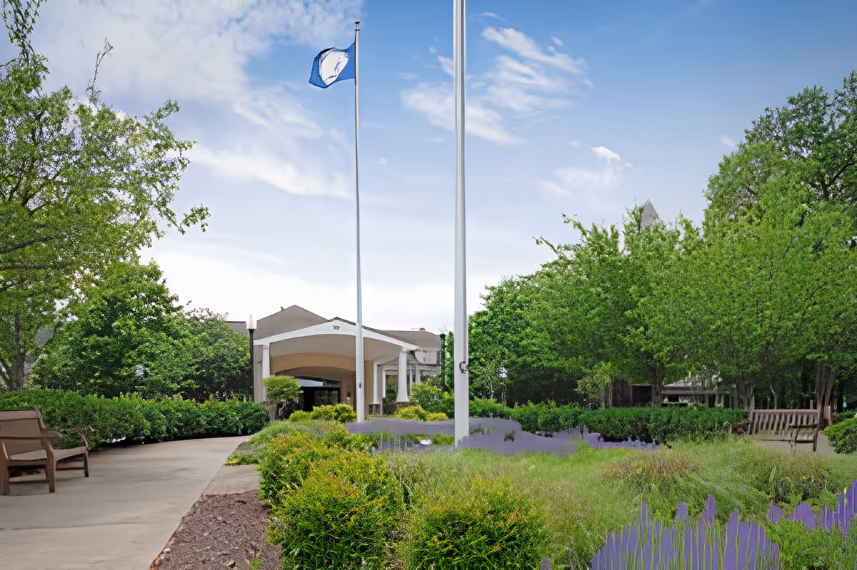 Outdoor view of the entrance to Belvoir Woods Health Care Center with a pathway, benches, green bushes, trees, and two flagpoles, one with a blue flag waving under a partly cloudy sky.