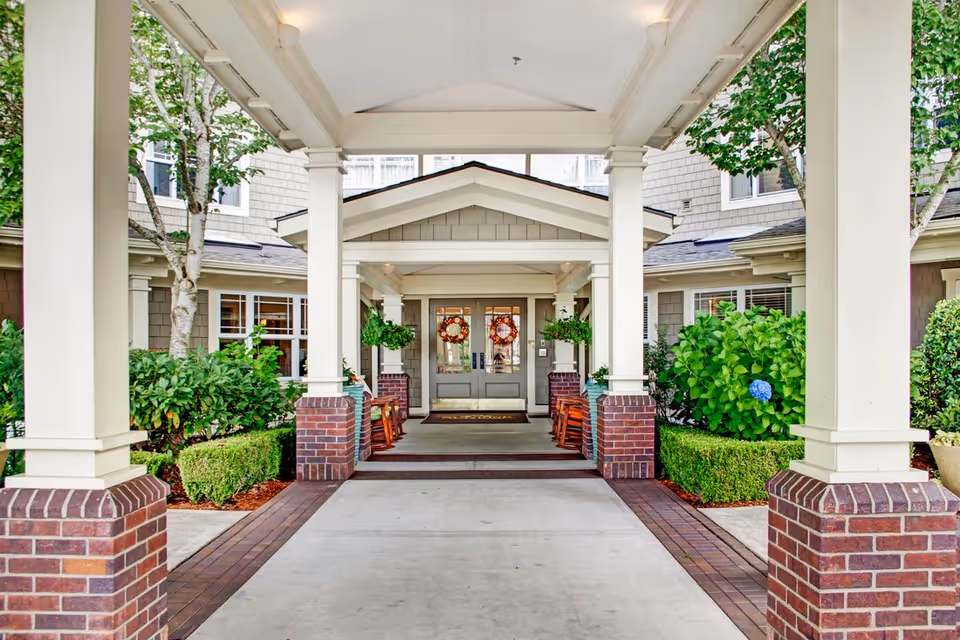 Covered entrance of a senior living facility with white columns on brick bases, green shrubs and trees on both sides, and double doors decorated with wreaths at the end of the walkway.