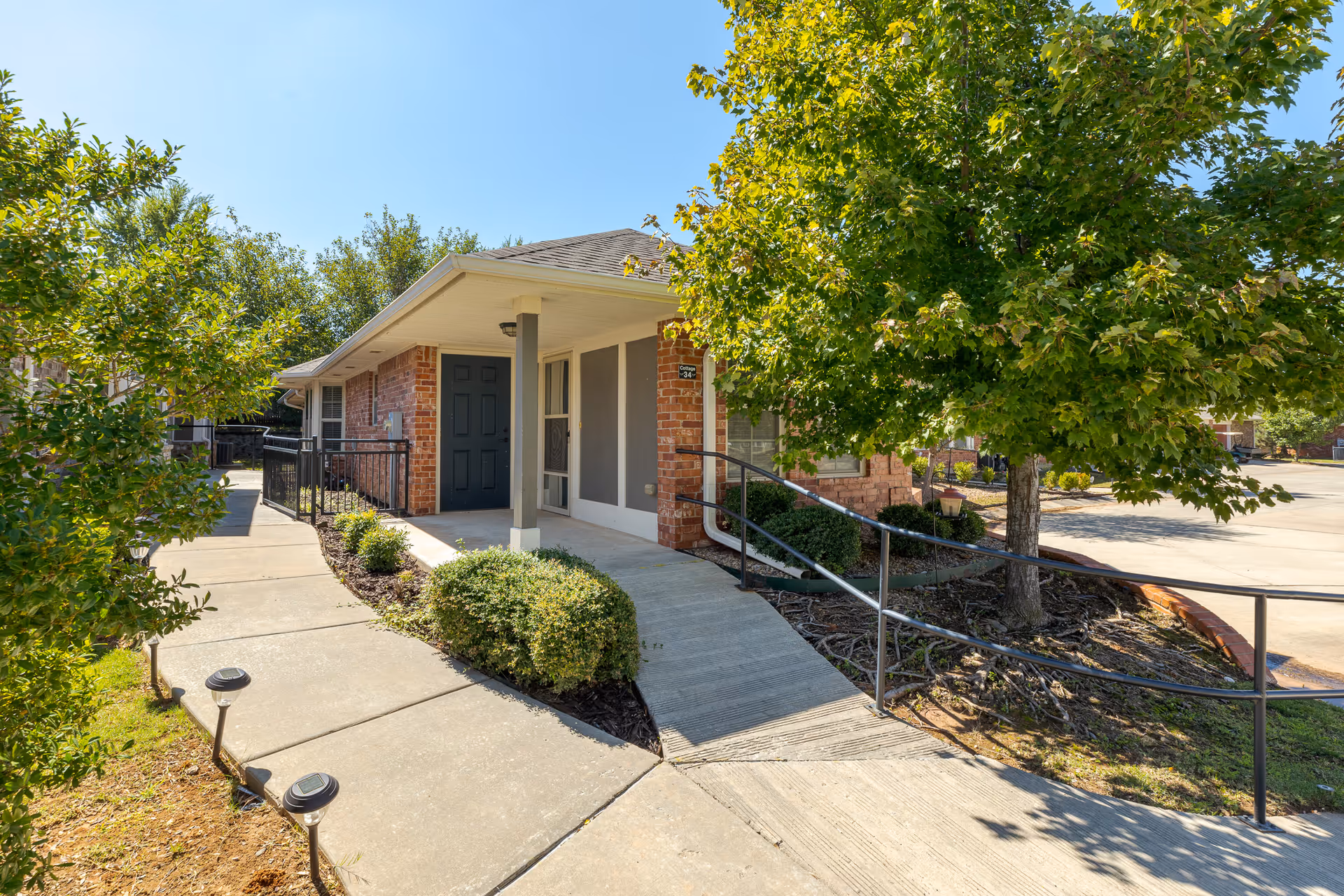 Exterior view of a single-story brick building with a covered entrance, a black door, and a ramp with handrails leading up to the entrance. The surrounding area features a concrete walkway, green bushes, a tree, and clear blue sky.