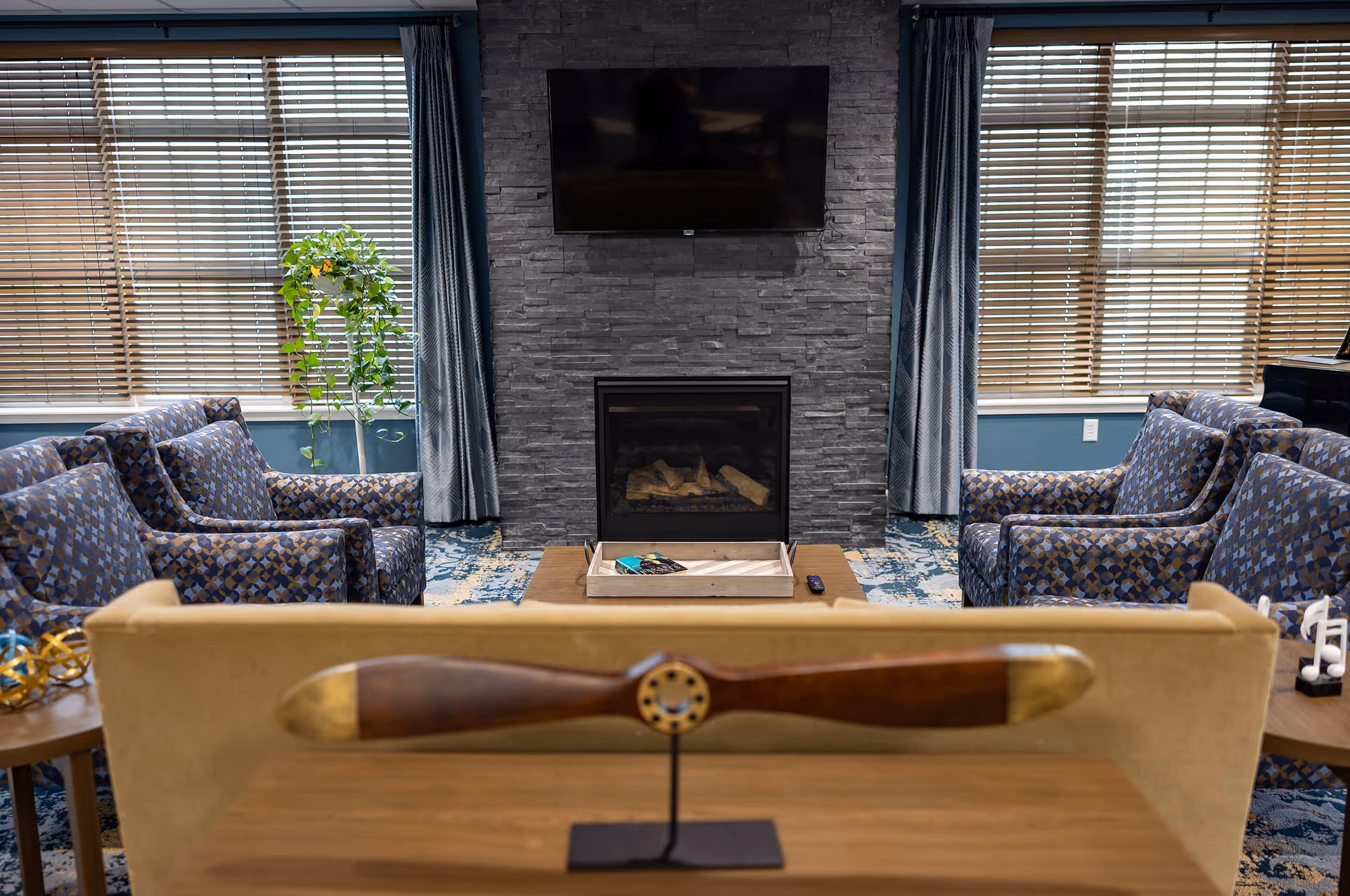 A cozy living room with a stone fireplace and a mounted flat-screen TV above it. The room features two patterned armchairs on each side, a wooden coffee table with a tray and remote control, and a beige sofa in the foreground with a decorative wooden propeller on a stand. Large windows with wooden blinds and blue curtains flank the fireplace, and a green potted plant is visible near the left window.