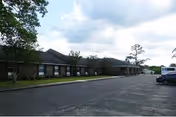 Front exterior of a single-story brick senior care building with a parking lot, trees, and a cloudy sky.