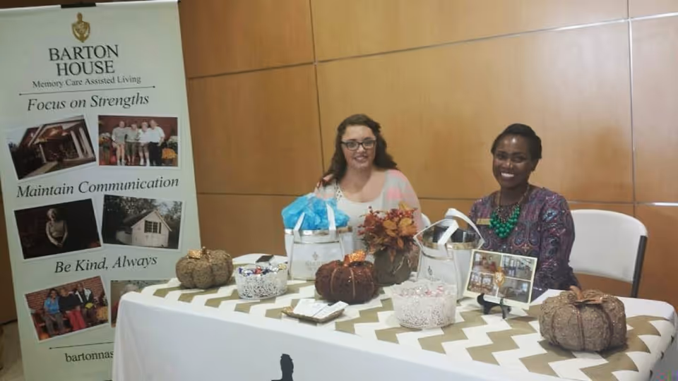 Two women sitting behind a decorated table with fall-themed items including decorative pumpkins, a small bouquet of autumn leaves, and baskets with candy. Behind them is a banner for Barton House Memory Care Assisted Living with phrases like 'Focus on Strengths,' 'Maintain Communication,' and 'Be Kind, Always.' The setting appears to be an indoor event or informational booth.