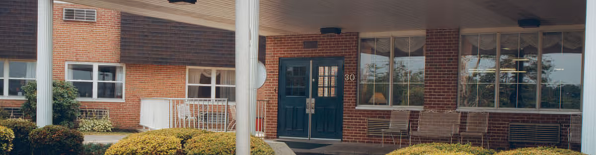 Entrance of a brick building with large windows and double blue doors under a covered porch, surrounded by trimmed bushes and greenery.