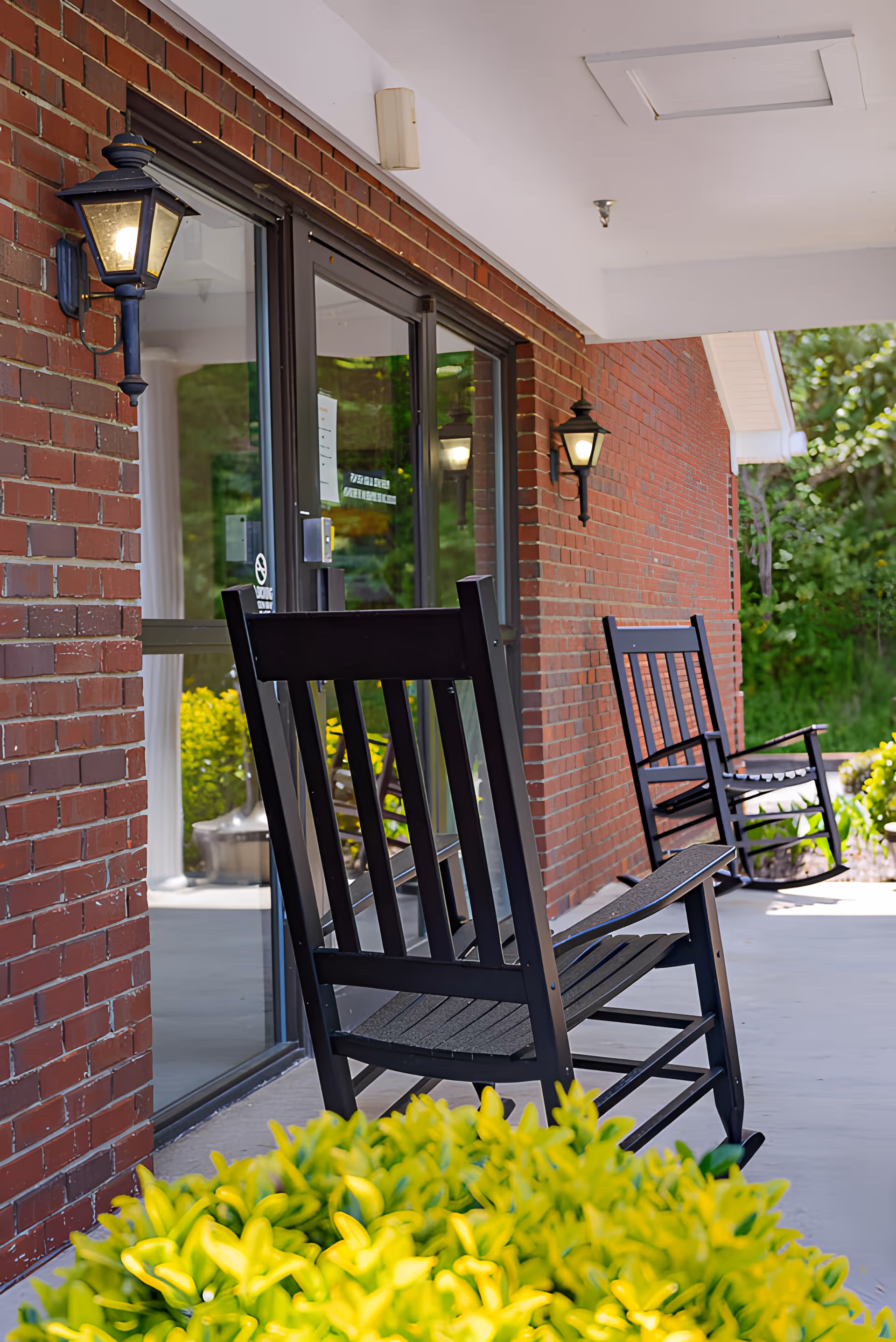 Outdoor porch area of a brick building with two black rocking chairs on a concrete floor, wall-mounted lantern-style lights, and green shrubbery in the foreground.