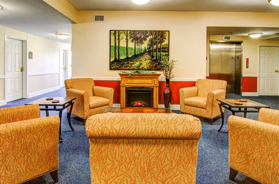 A cozy senior living community lounge area with four patterned armchairs arranged around a wooden coffee table and two side tables. A wooden electric fireplace with a decorative plant on top is centered against the wall, above which hangs a framed painting of a tree-lined path. The room has blue carpet, cream-colored walls with a red accent panel, and an elevator and doors visible in the background.