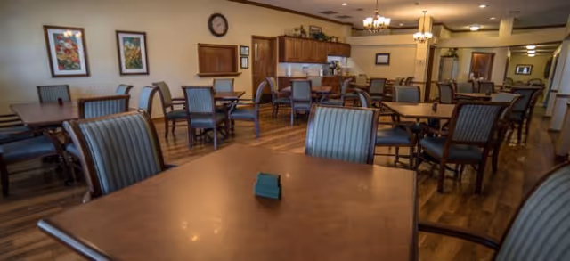 Spacious communal dining room with multiple wooden tables and upholstered chairs, a serving counter and warm overhead lighting.