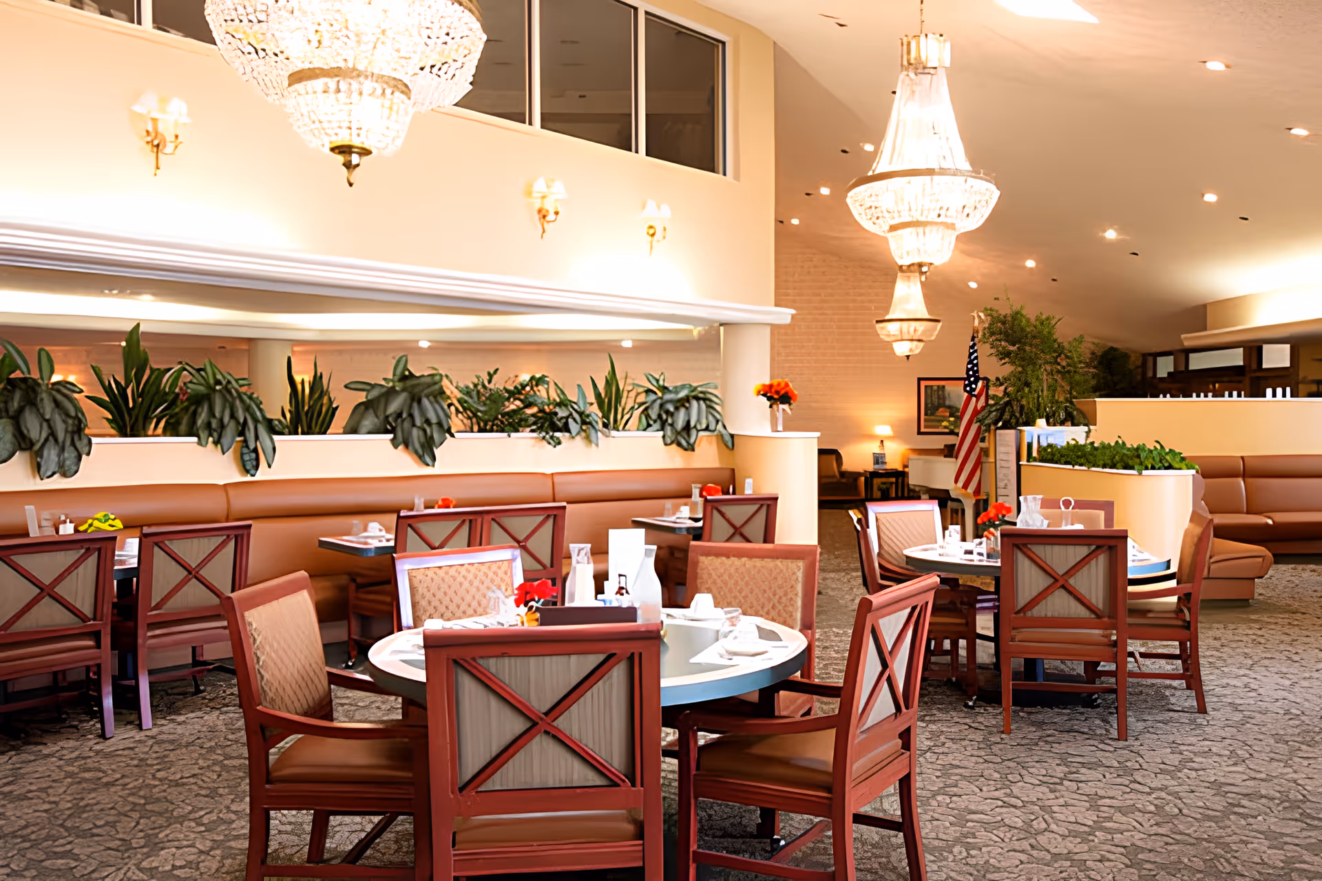 A well-lit dining room with round tables and wooden chairs arranged neatly. The room features large chandeliers, potted plants, and an American flag in the background. The walls are light-colored with sconces, and there is a carpeted floor.