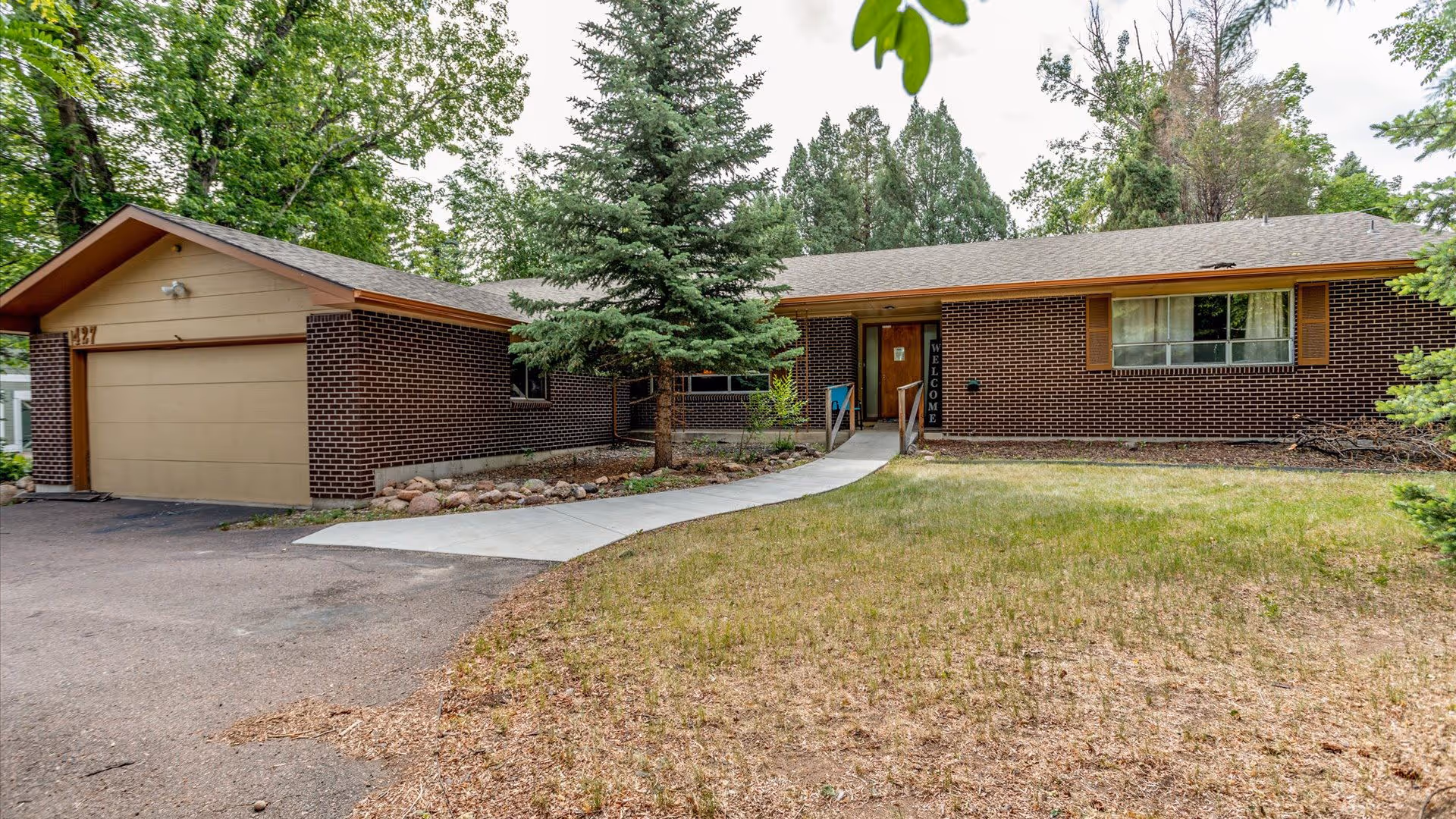 Front exterior view of a single-story brick building with a two-car garage on the left and a main entrance with a 'WELCOME' sign on the right. The building is surrounded by trees and has a concrete walkway leading to the entrance.