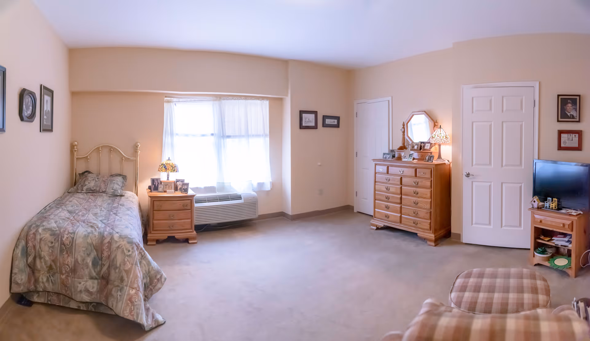 A senior living bedroom with a single bed covered in a patterned quilt, a wooden nightstand with a lamp and framed photos, a window with sheer curtains, a wooden dresser with a mirror and lamp, a TV on a small wooden stand, and a plaid armchair in the foreground. The walls are light beige and decorated with framed pictures.