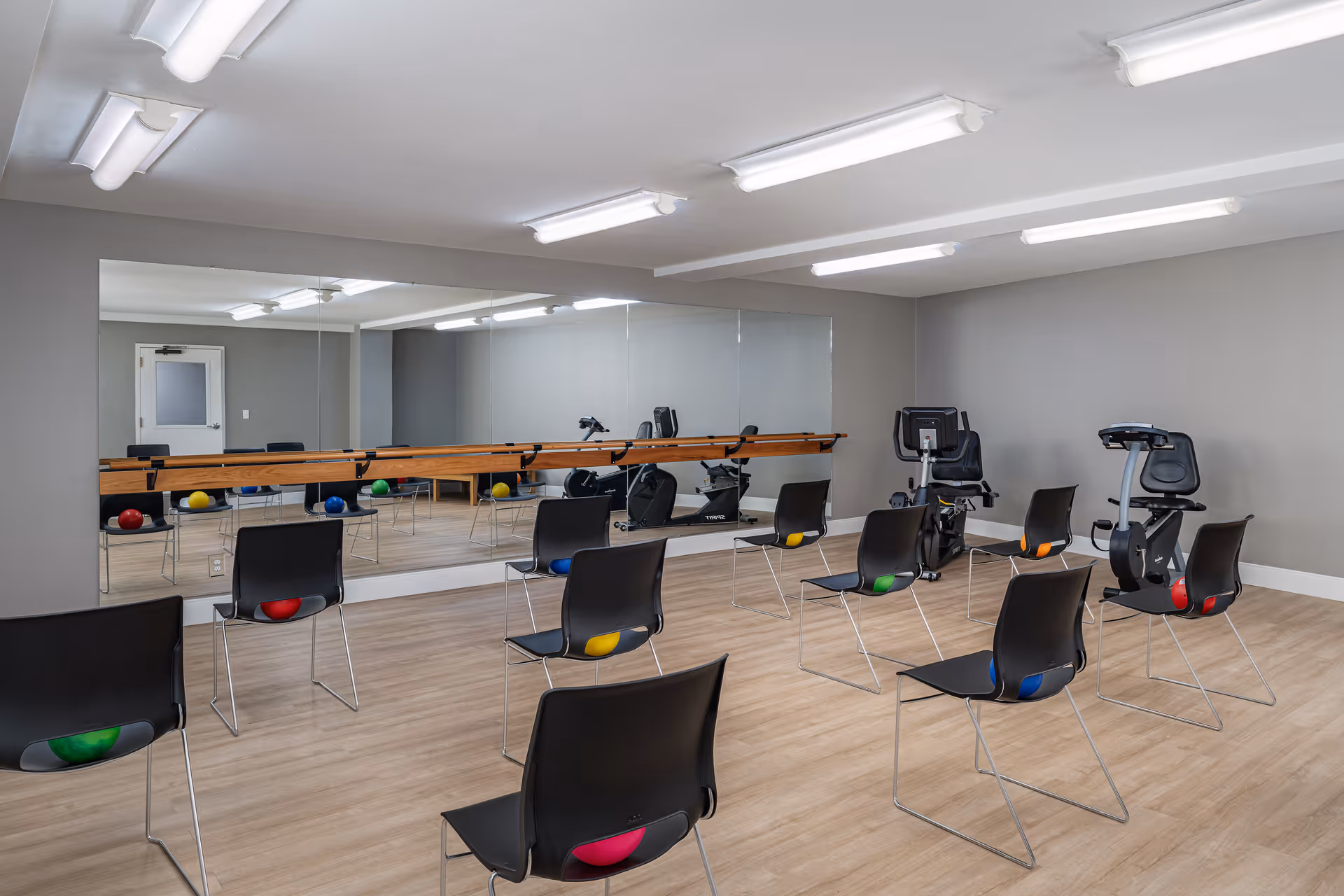 A fitness room with several black chairs arranged in rows, each with a colorful exercise ball placed underneath. Two stationary exercise bikes are positioned against the right wall. A large mirror covers the left wall, reflecting the room and equipment. The room has wood flooring, gray walls, and bright overhead fluorescent lighting.