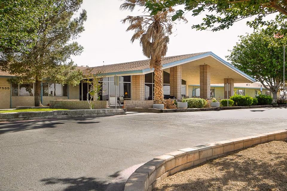 Front exterior of a single-story convalescent hospital with a covered entrance, driveway, palm tree, and landscaped grounds.