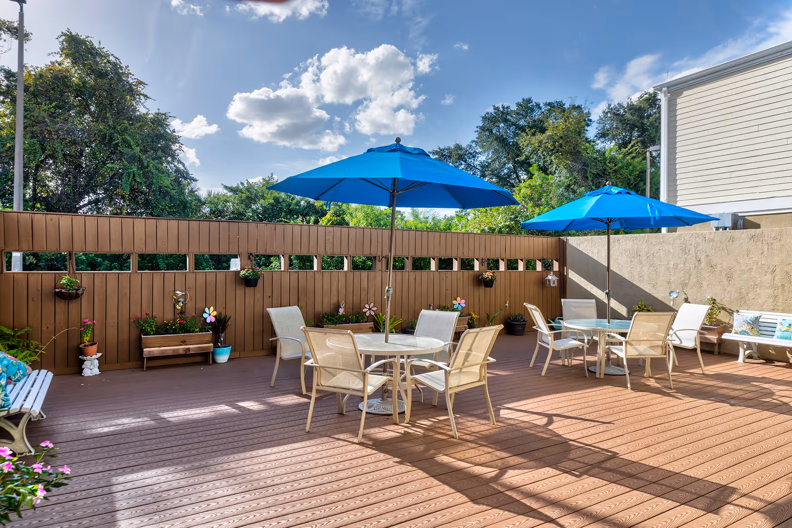 A spacious outdoor patio featuring blue umbrellas, white tables, and chairs surrounded by greenery and colorful flower pots.