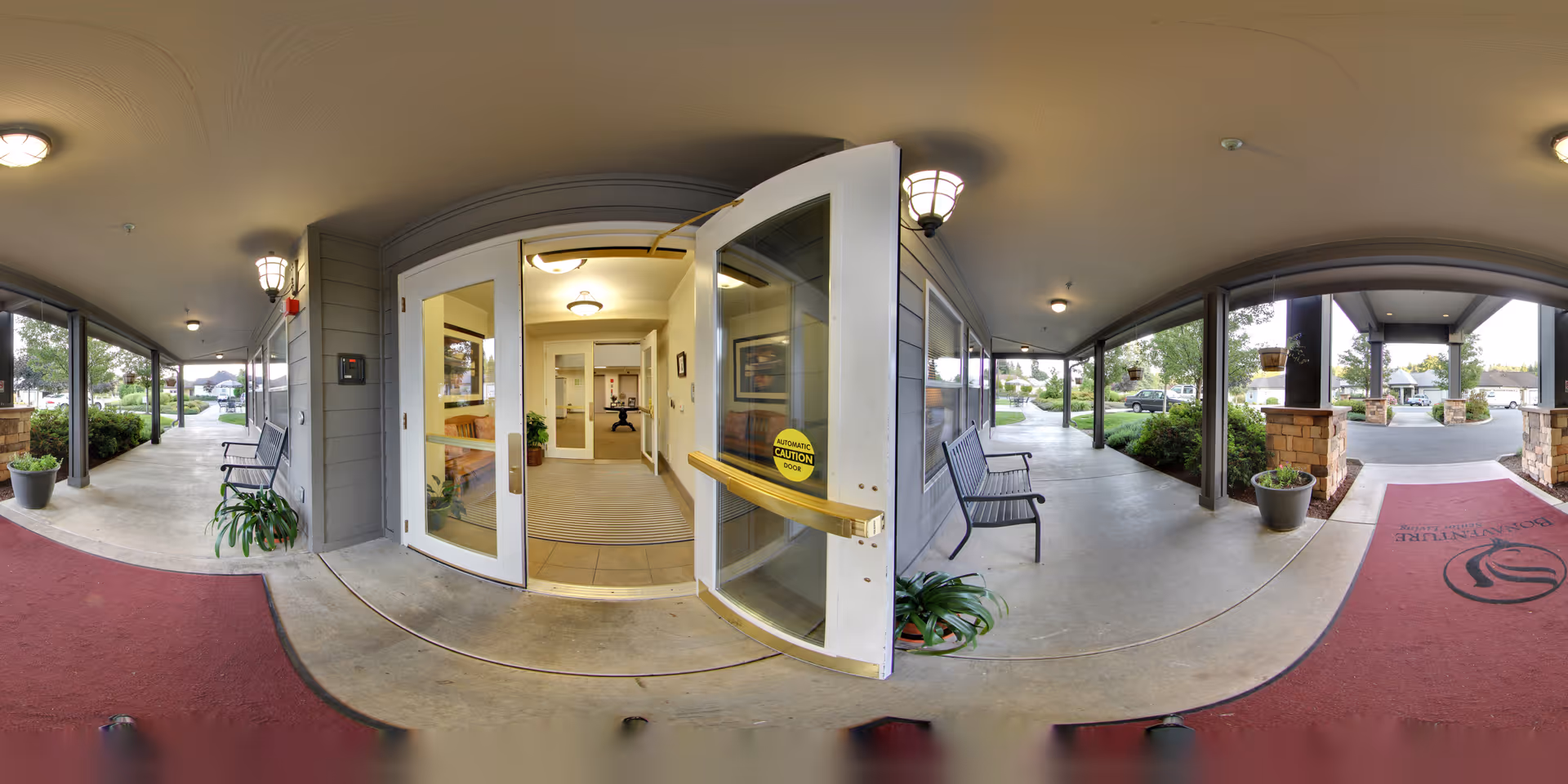 Covered entrance with an open glass door into a lobby, benches and potted plants along the walkway and a red entrance mat.