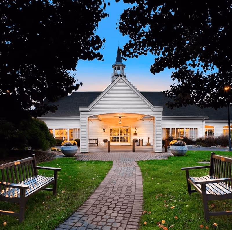 Front exterior view of a white building with a peaked roof and a small cupola on top, illuminated warmly at dusk. A brick pathway leads to the entrance, flanked by green lawns and wooden benches on either side. Trees frame the scene from above.