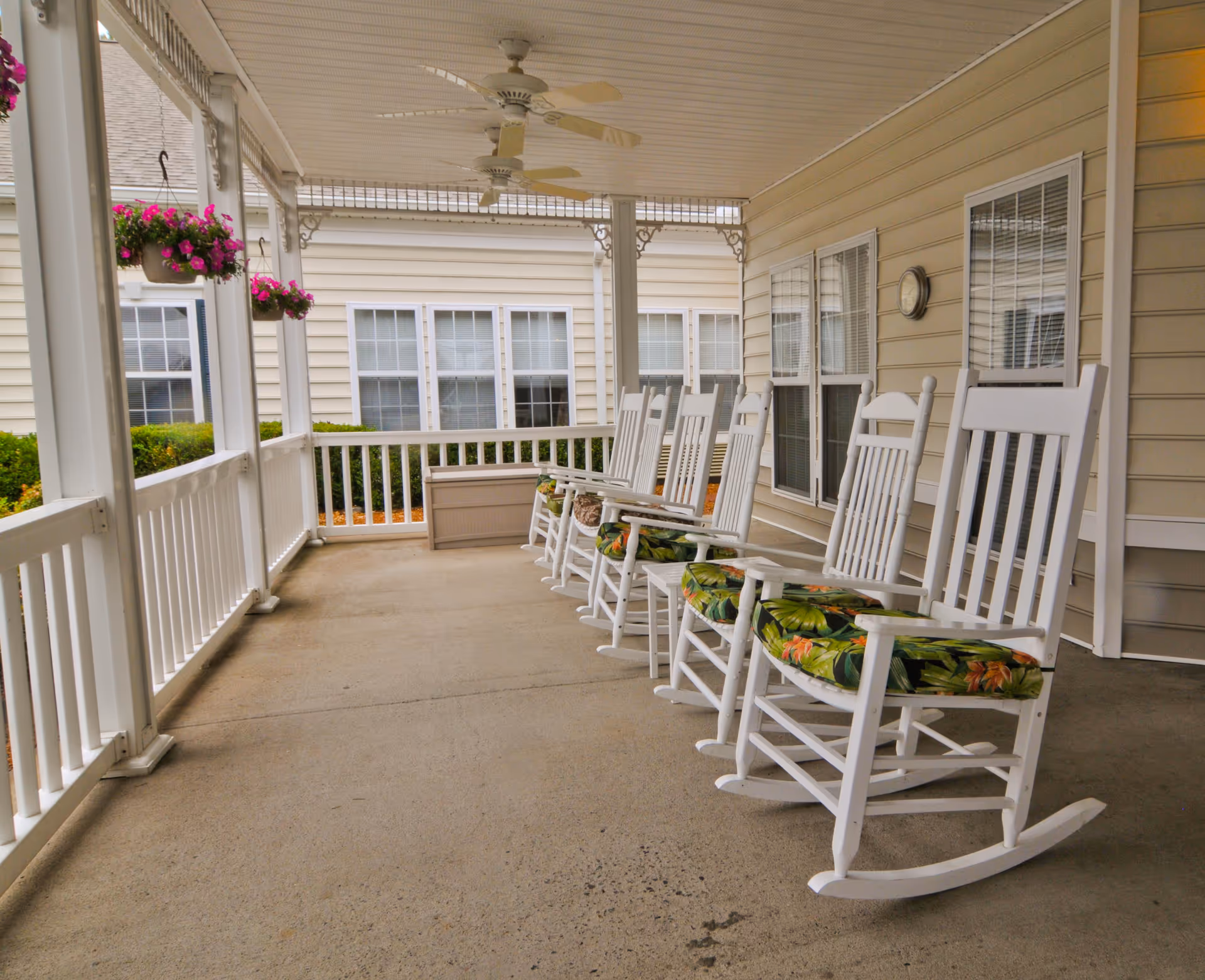 Covered porch area with six white wooden rocking chairs with floral cushions lined up facing outward. The porch has white railings, ceiling fans, hanging flower pots with pink flowers, and beige siding on the building walls with multiple windows.