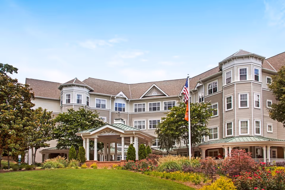 Exterior view of a large, multi-story senior living facility with beige siding, multiple windows, and a covered entrance. The building is surrounded by well-maintained landscaping including trees, shrubs, and colorful flowers. An American flag and another flag are displayed on a flagpole in front of the building under a clear blue sky.