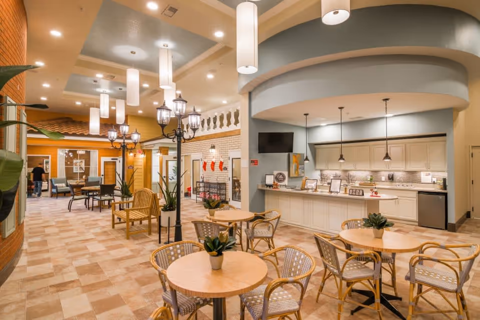 Interior view of a senior living facility common area with round tables and chairs, potted plants on tables, a kitchen counter with pendant lights, and decorative street lamp-style lighting. The space has a warm and inviting atmosphere with a mix of modern and classic design elements.