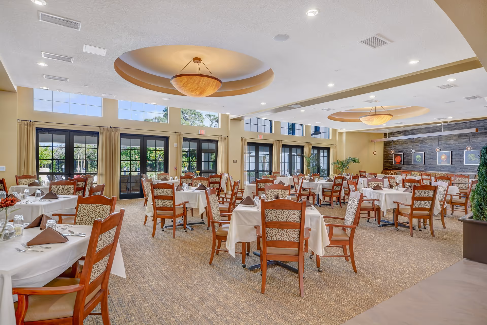 A spacious dining room with multiple tables covered in white tablecloths and set with brown napkins, glasses, and silverware. The room features large windows and glass doors letting in natural light, beige walls, carpeted floor, and decorative ceiling lights. There are framed pictures of vegetables on a stone accent wall and some potted plants in the corners.