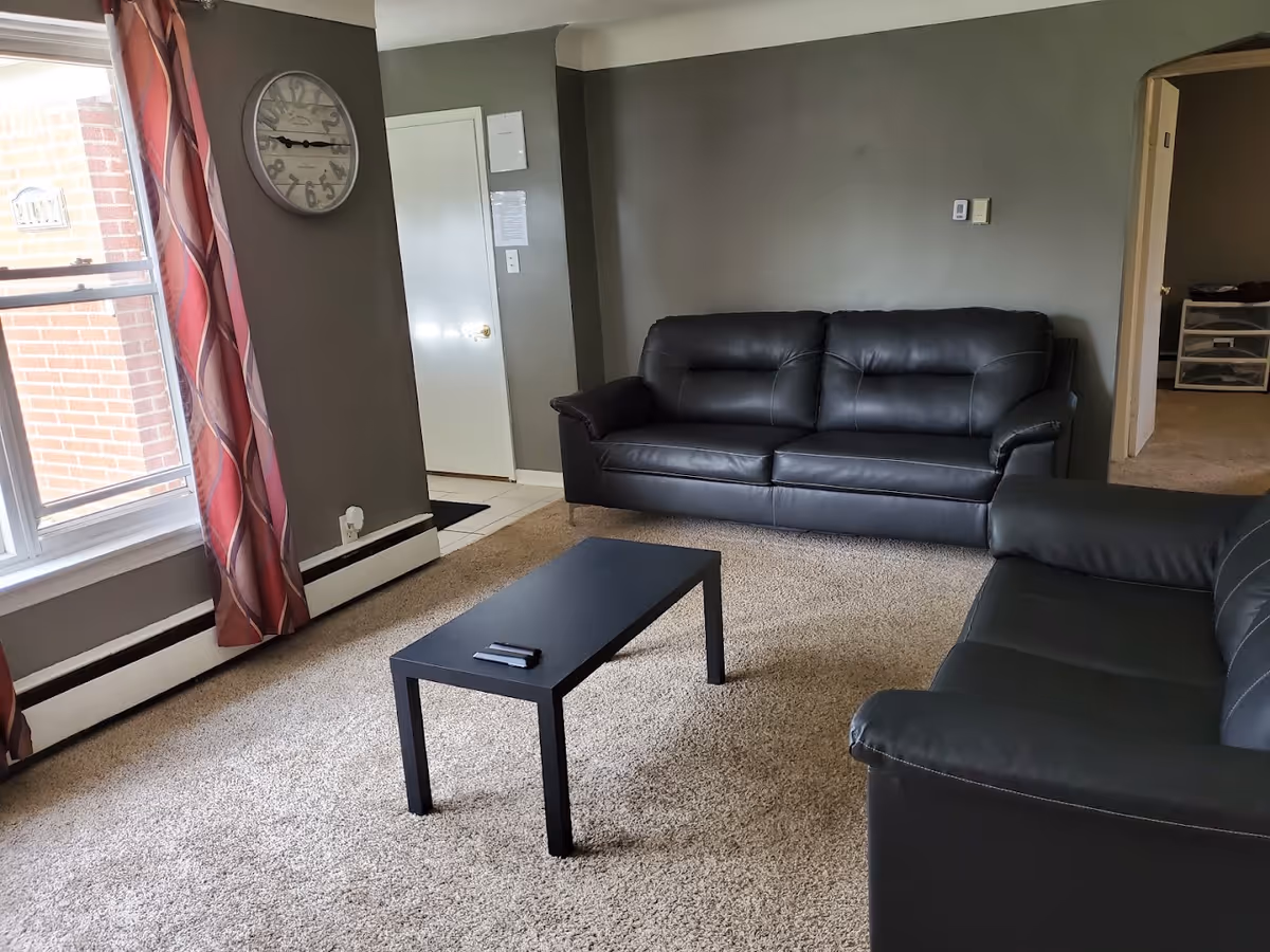 A living room with two black leather sofas facing each other, a small black coffee table with two remote controls on it, beige carpet flooring, gray walls, a large window with red and pink patterned curtains, and a round wall clock above a white door.