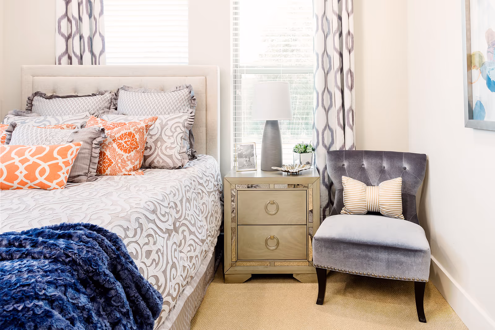 A cozy bedroom featuring a bed with multiple decorative pillows in shades of gray and orange, a patterned bedspread, a blue throw blanket, a mirrored nightstand with a lamp, a small plant, and a framed photo, next to a gray upholstered chair with a striped pillow, all set against a backdrop of windows with patterned curtains.
