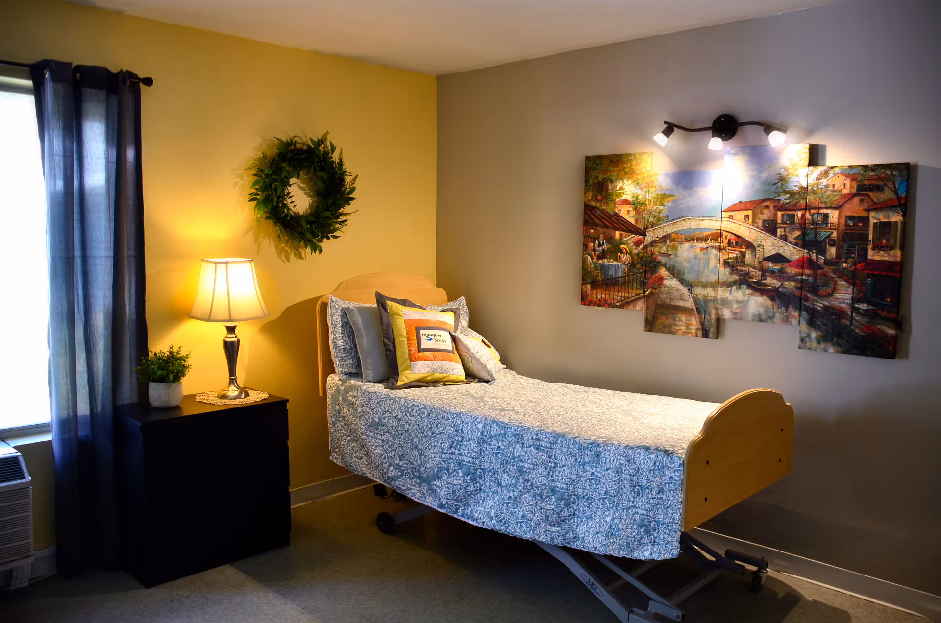 A single bed with a blue patterned bedspread and multiple pillows in a room with yellow and gray walls. A black nightstand with a lamp and a small plant is next to the bed. A green wreath hangs on the yellow wall above the nightstand, and a multi-panel painting of a scenic canal with buildings and a bridge is mounted on the gray wall above the bed. A window with dark curtains is on the left side of the room.