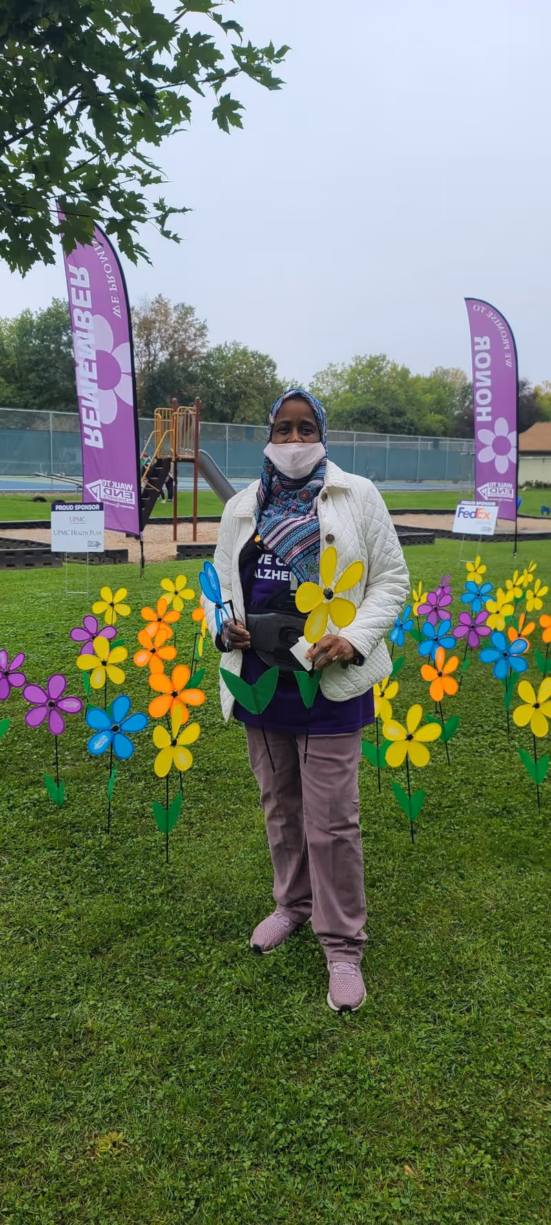 A person wearing a headscarf, face mask, white jacket, and purple shirt stands outdoors on grass surrounded by colorful pinwheel flowers. Behind them are two purple flags with text and logos, a playground slide, and a fenced area with trees in the background.