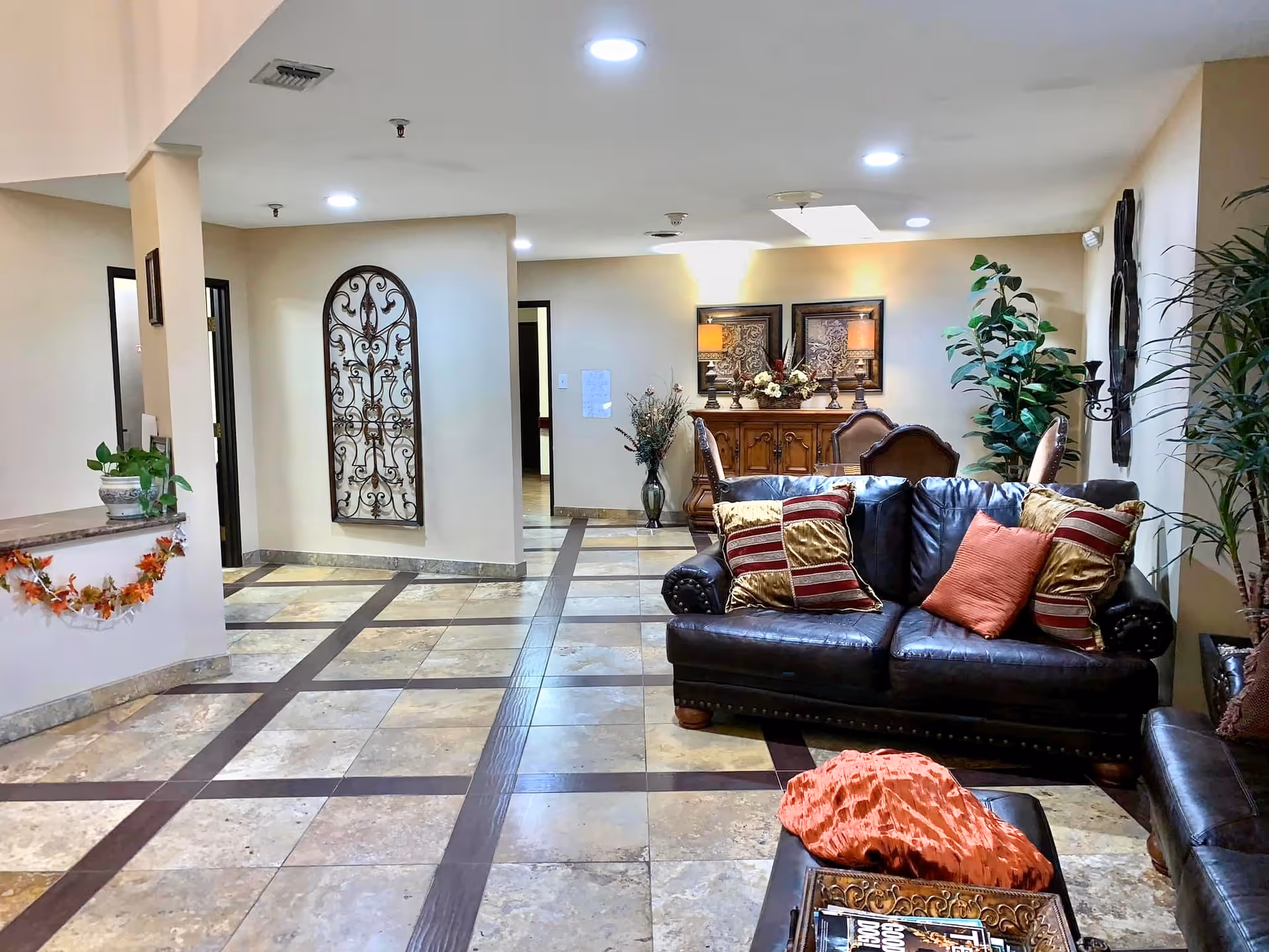 Interior view of a senior living facility lounge area with tiled floors, a dark leather sofa adorned with decorative pillows, a coffee table with a tray and magazines, potted plants, wall art, and a wooden sideboard with lamps and floral arrangements in the background.