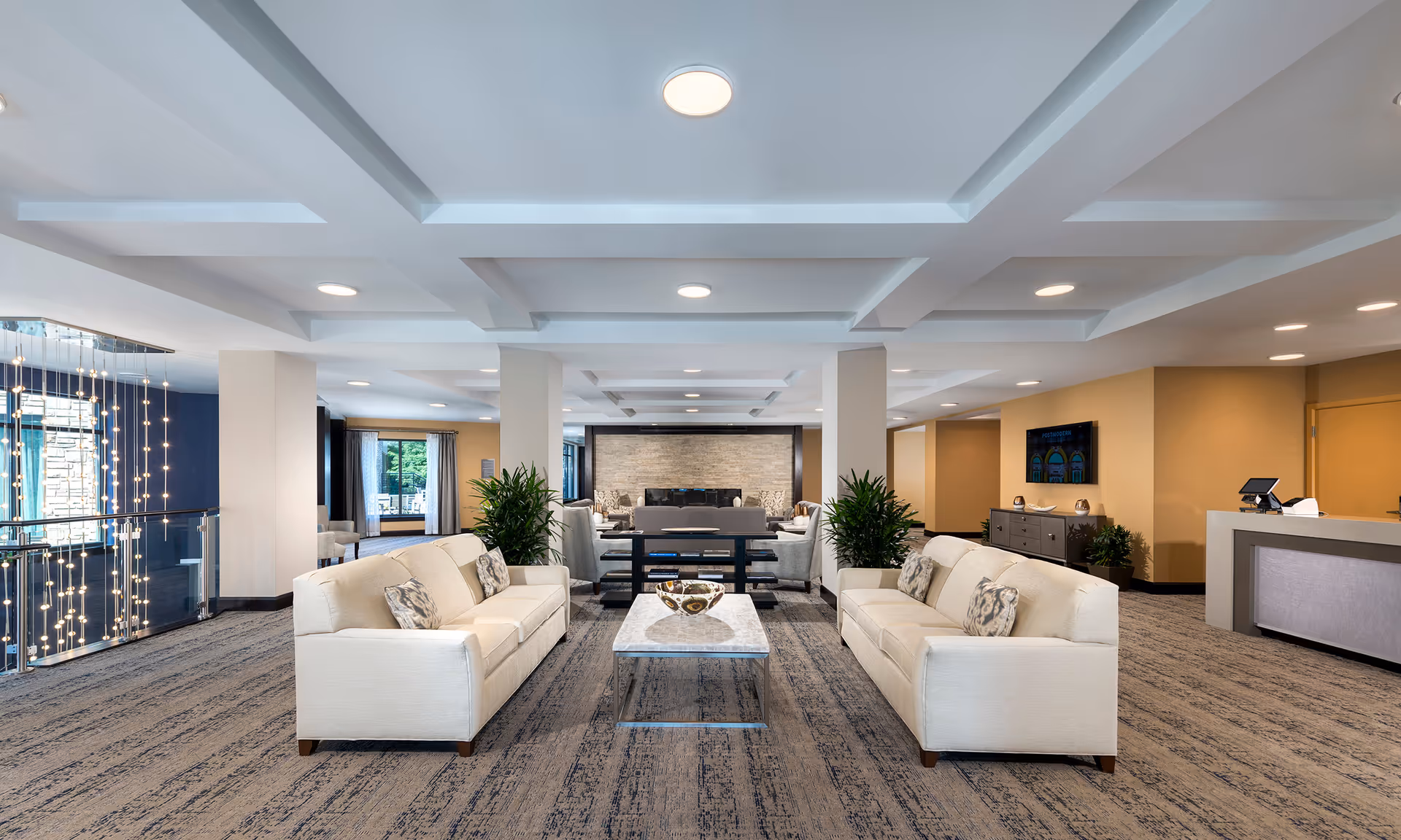 A spacious and modern common area with two white sofas facing each other across a marble coffee table. The room features a coffered ceiling with recessed lighting, carpeted floor, potted plants, and a reception desk on the right side. Large windows and glass railings with hanging lights are visible on the left side.