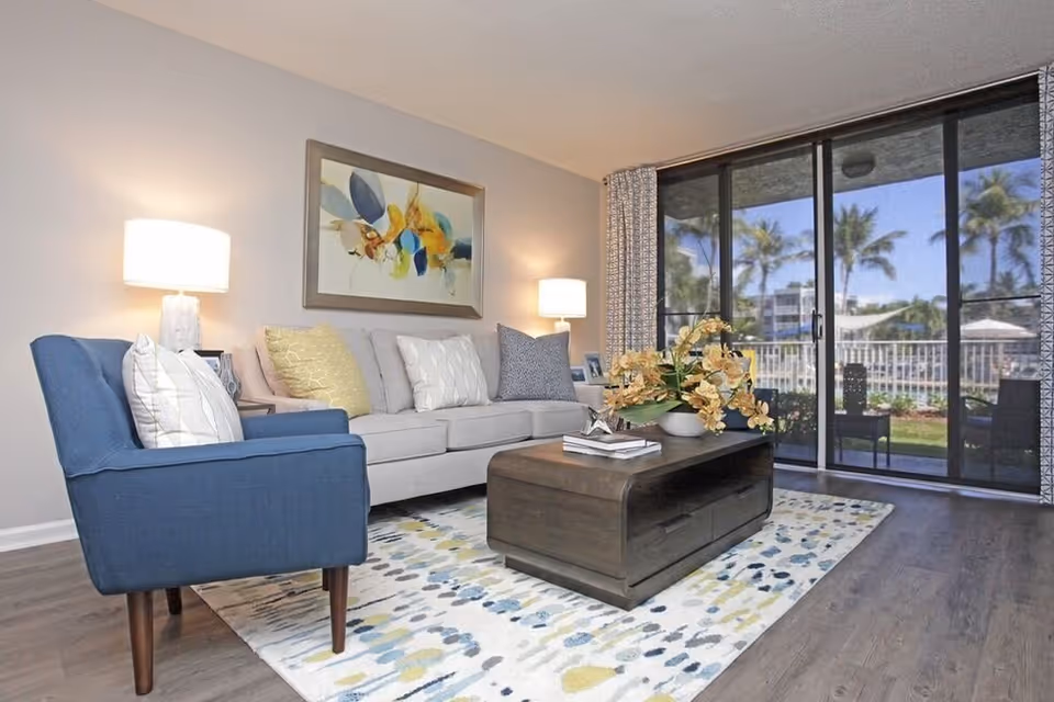 A modern living room with a blue armchair and a light gray sofa adorned with multiple cushions. A wooden coffee table with a flower arrangement and books sits on a patterned rug. Two table lamps provide warm lighting, and large sliding glass doors reveal an outdoor view with palm trees and buildings.