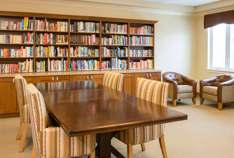 A cozy reading room with a large wooden table surrounded by six striped upholstered chairs. Behind the table is a built-in wooden bookshelf filled with books. To the right, there are two patterned armchairs near a window with a brown valance, allowing natural light into the room.