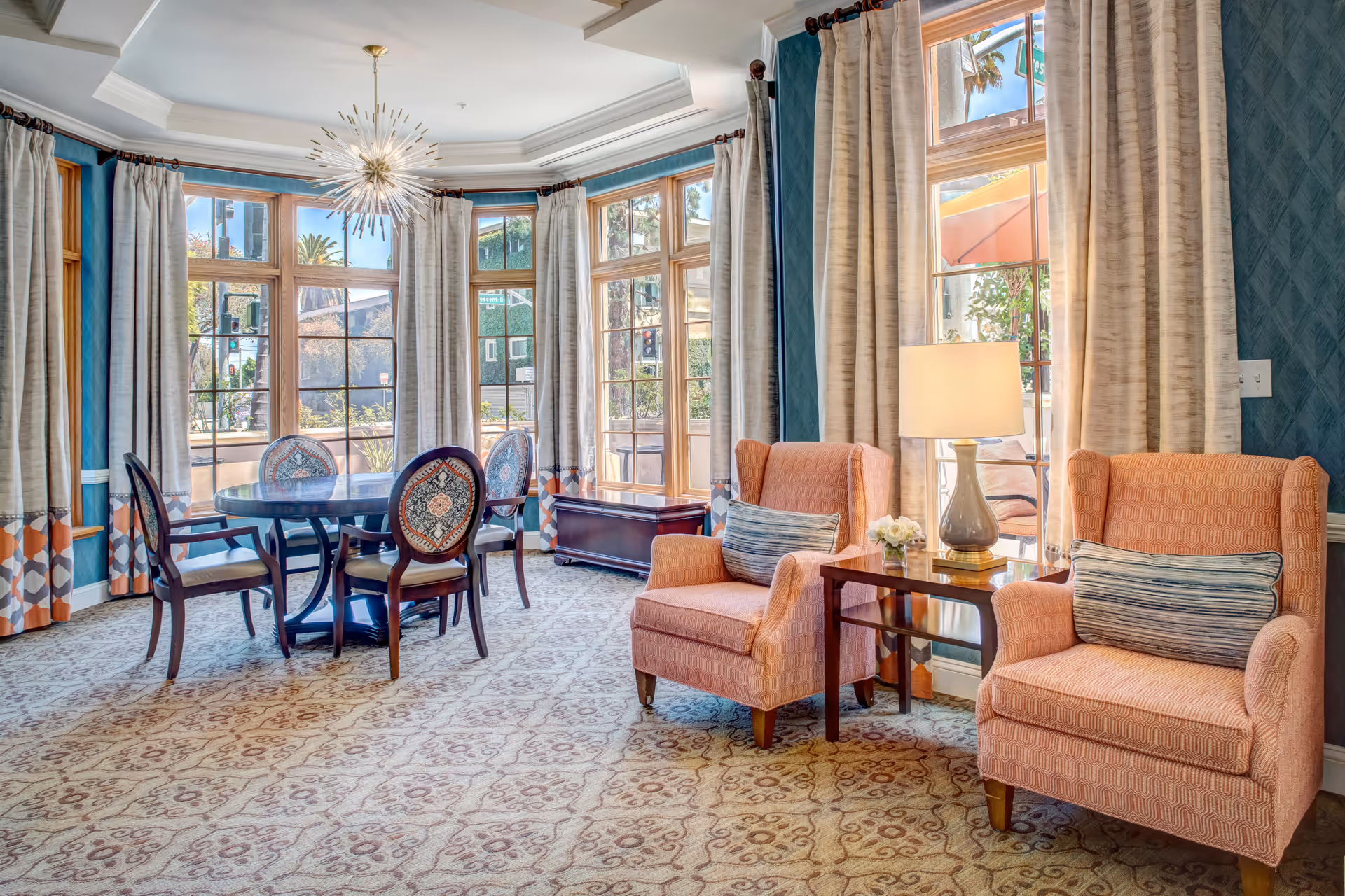 Bright communal sitting and dining area with a round table by tall bay windows and two upholstered armchairs with a side table and lamp.
