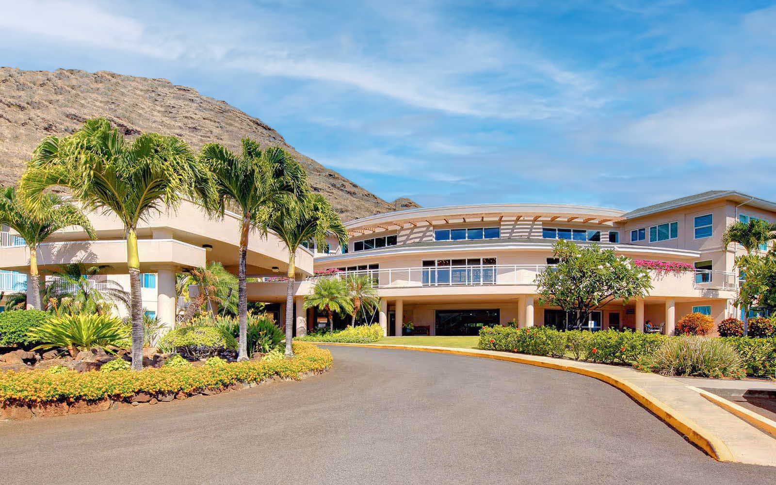 Front entrance and driveway of a tropical senior living building with palm trees, landscaping, and a mountain backdrop.