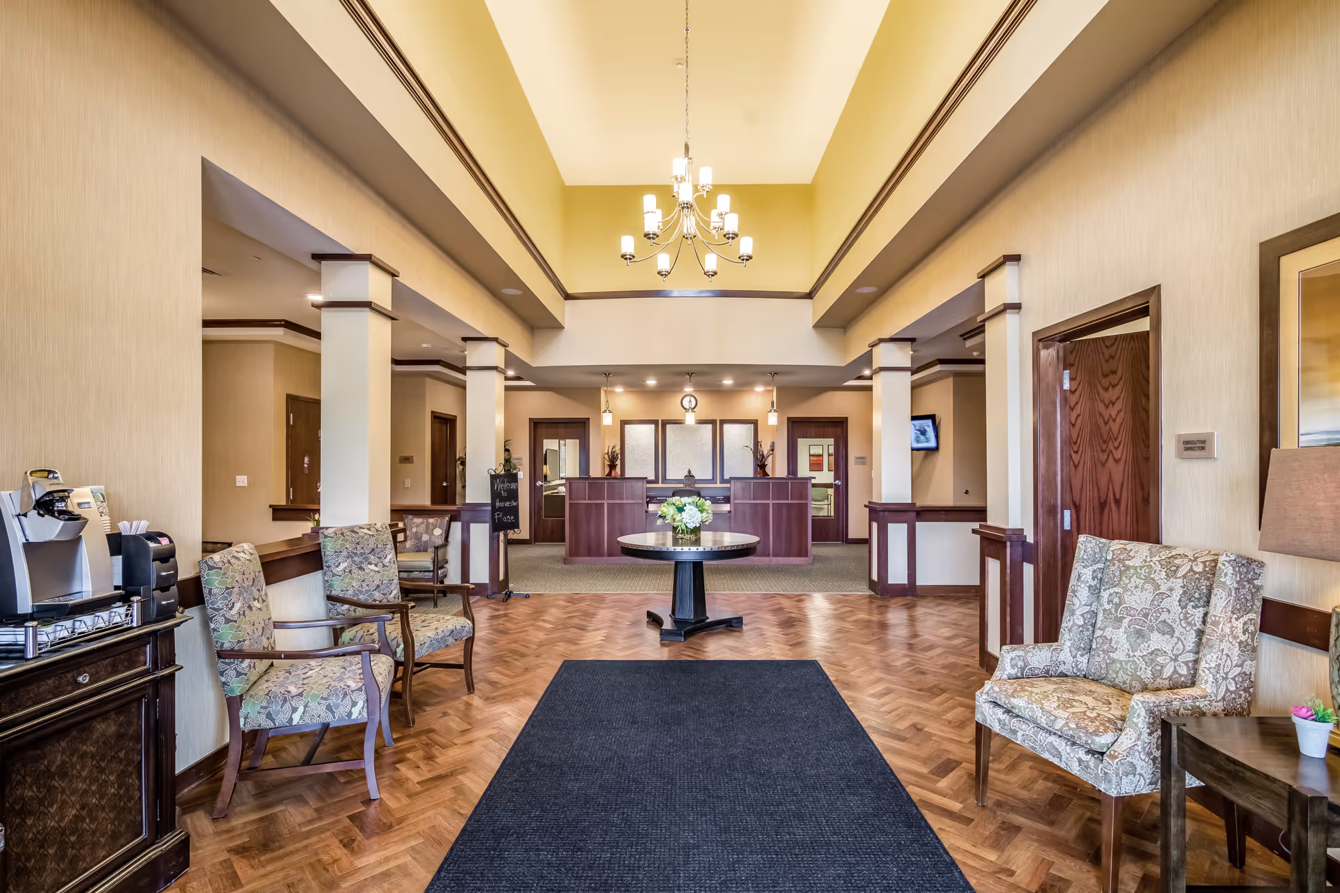 A spacious and well-lit reception area with a central round table holding a flower arrangement. The room features a high ceiling with a chandelier, patterned armchairs along the sides, a coffee machine on a cabinet to the left, and a sign that reads 'Welcome to Harvester Place'. The reception desk is visible in the background with warm lighting and wooden accents.