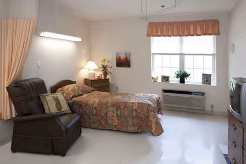 A furnished senior living bedroom with a bed, recliner, nightstand, window, and TV.